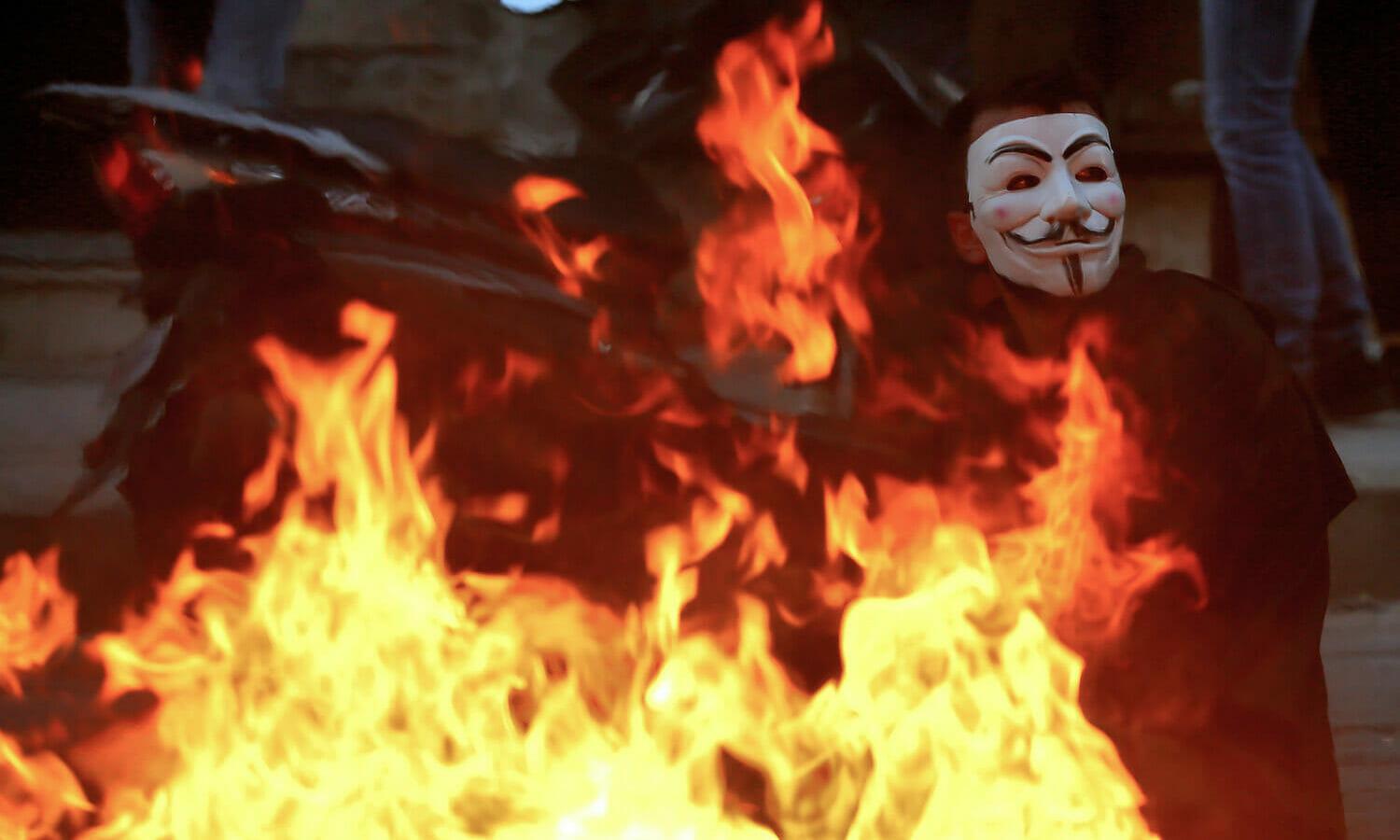 An anti-government activist, displaying anonymous mask, sits near bone fire during clashes with riot police in Beirut. (Marwan Naamani/picture alliance via Getty Images)