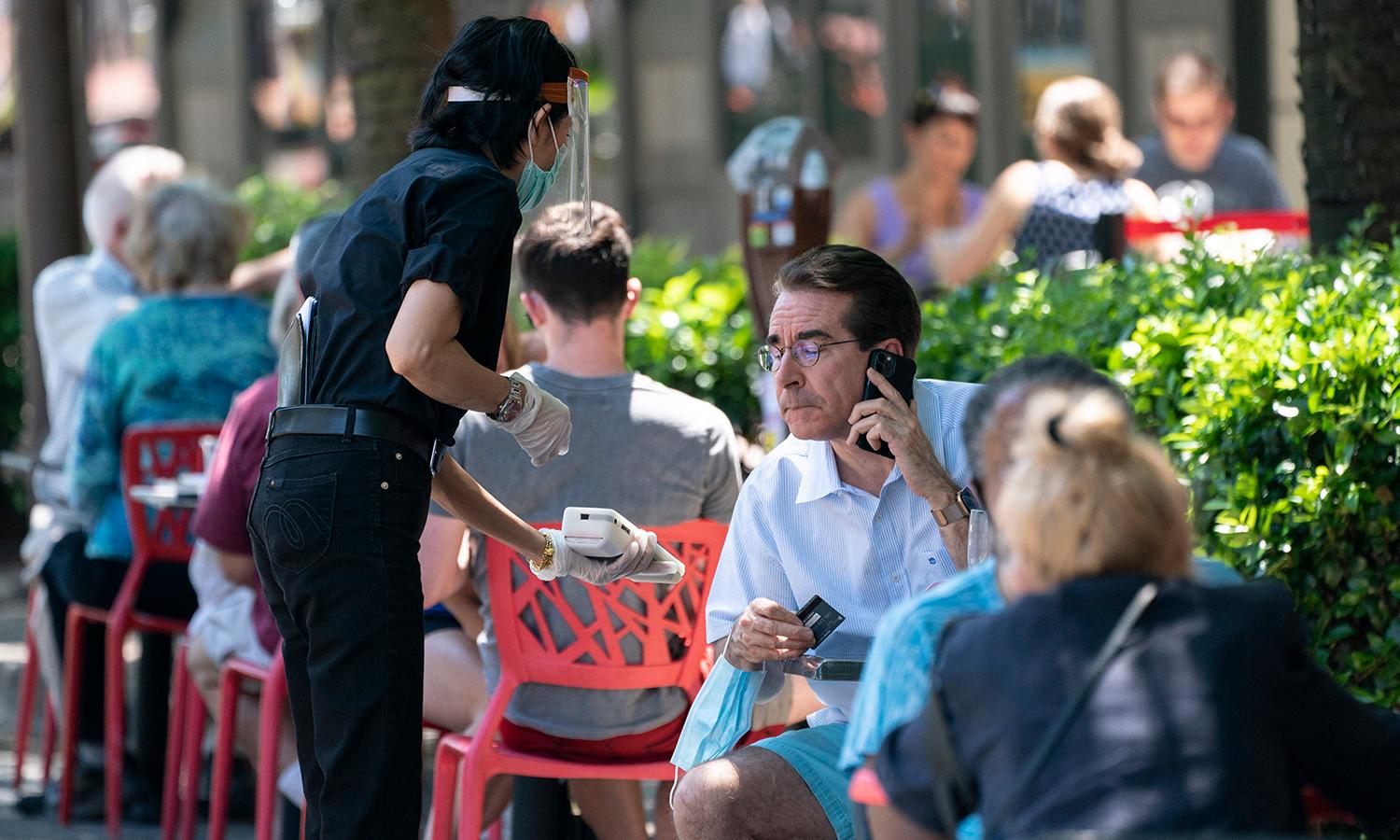 A waiter in wearing a protective face mask presents an outside diner a contactless payment system.