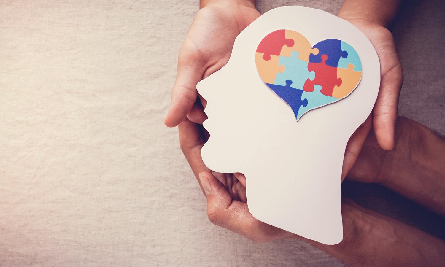 A silhouette of a person's head with a heart-shaped puzzle for a brain is held in the hands of two people.