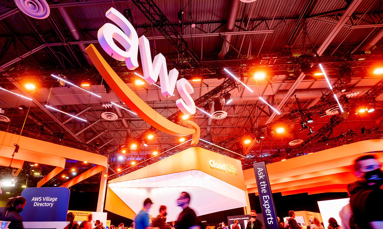 A logo for AWS is suspended over a convention room with attendees walking below.