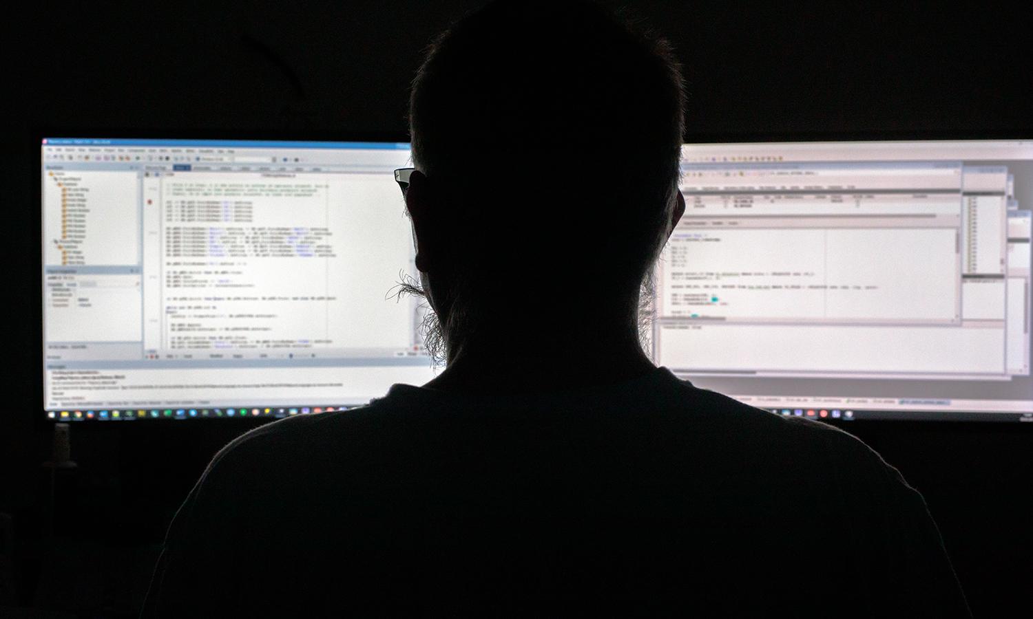 A silhouetted programmer sits in the dark in front of two computer monitors.