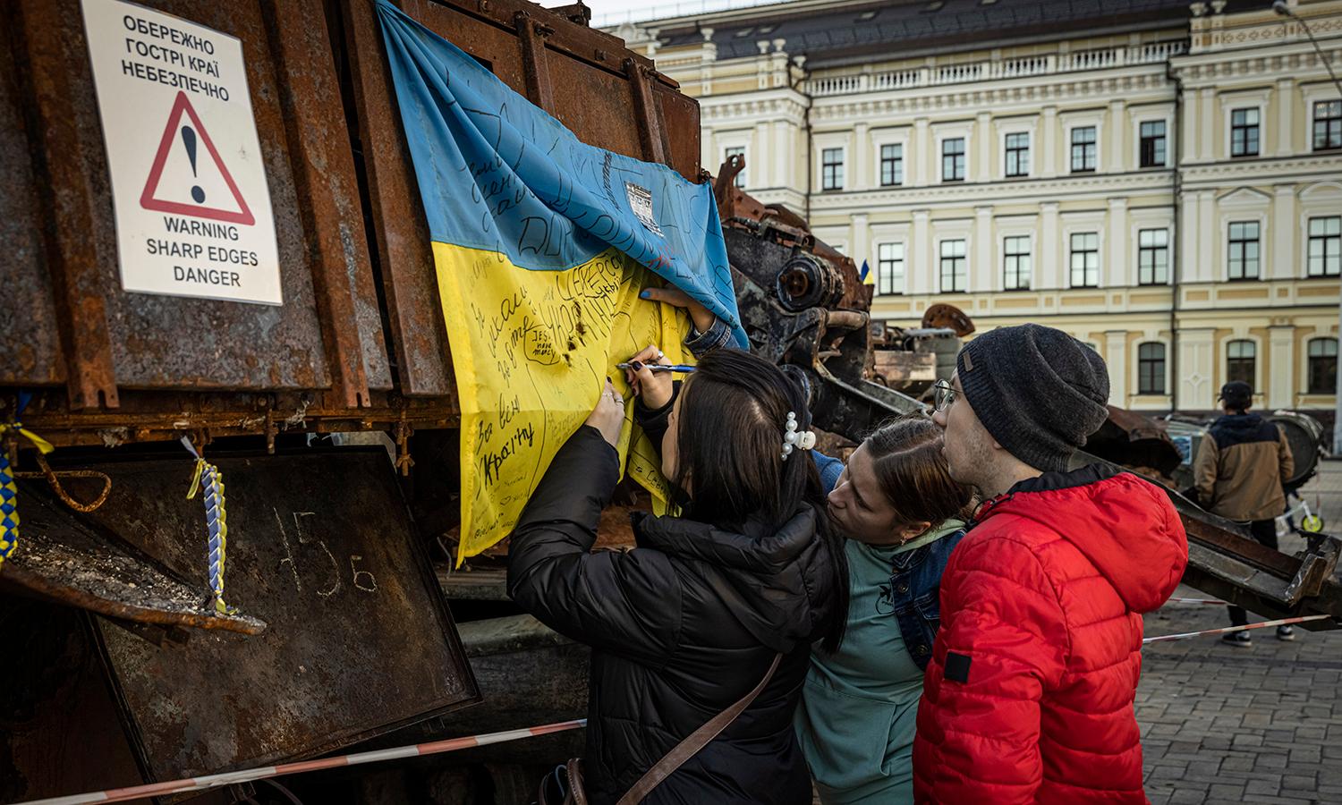 A girl writes one a Ukrainian flag as people look at an exhibition of tanks and military equipment on display.